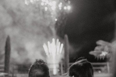 Un couple de dos regarde un feu d'artifice illuminer le ciel nocturne dans un cadre extérieur.