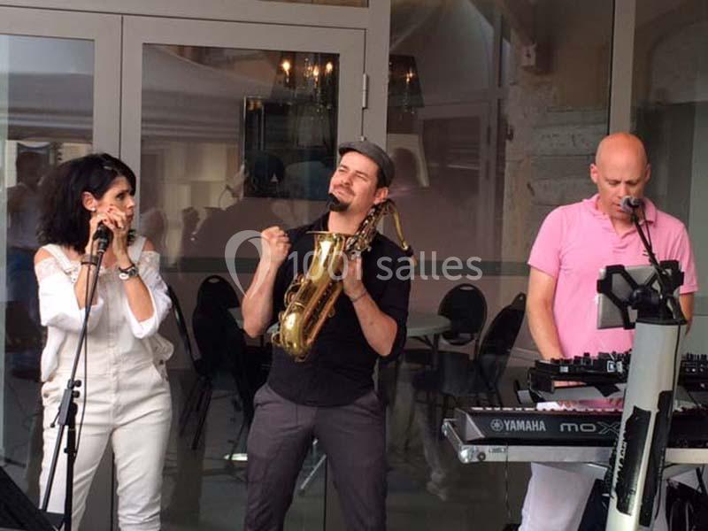 Trois musiciens jouent en extérieur, avec un saxophone, un clavier et un micro.