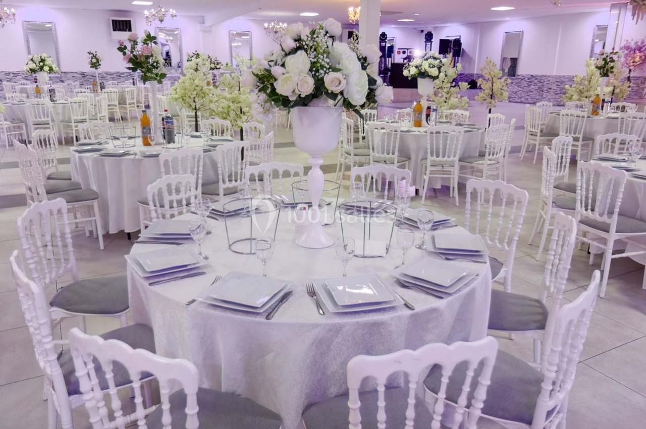 Salle de réception décorée avec des tables rondes, nappes blanches, chaises élégantes et centres de table fleuris.