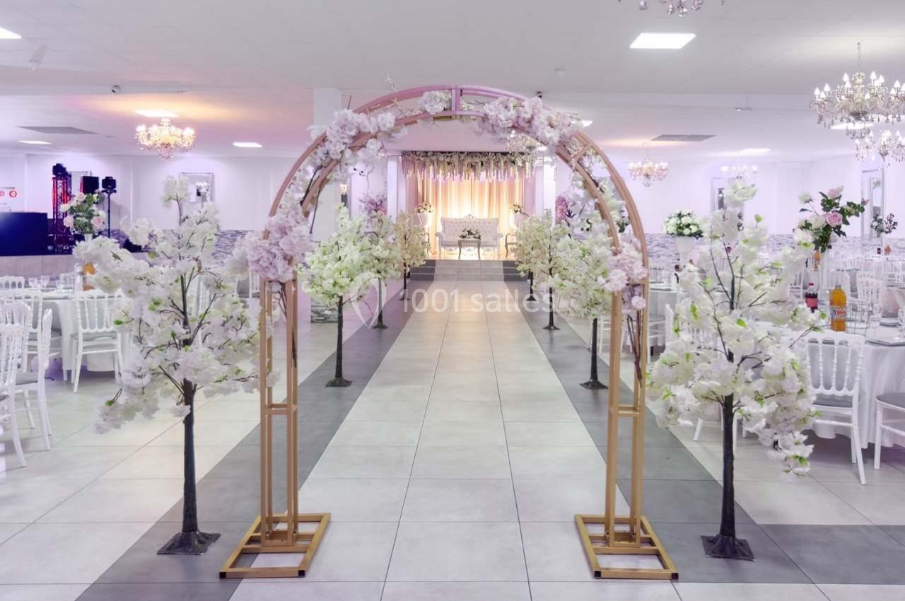 Arche décorée de fleurs blanches dans une salle de réception lumineuse avec des chaises et des tables alignées.