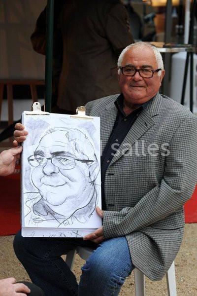 Un homme assis tenant un dessin caricatural de lui-même, réalisé sur une feuille attachée à une planchette.