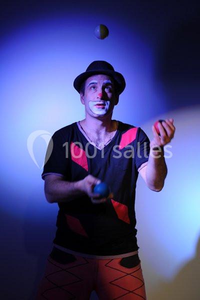 Un jongleur maquillé en lumière bleue jongle avec trois balles, portant un chapeau et des vêtements colorés.