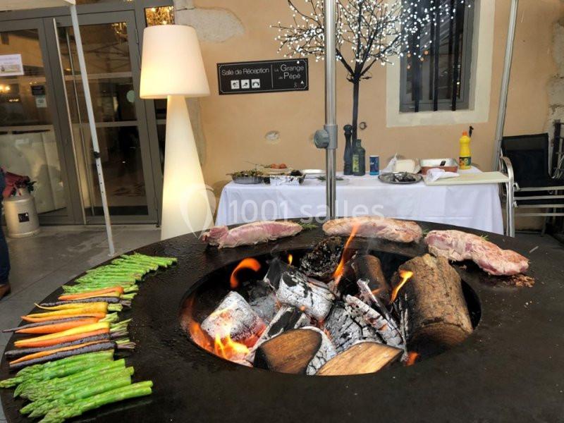 Légumes et viandes grillant sur un barbecue circulaire au feu de bois, avec une table dressée en arrière-plan.