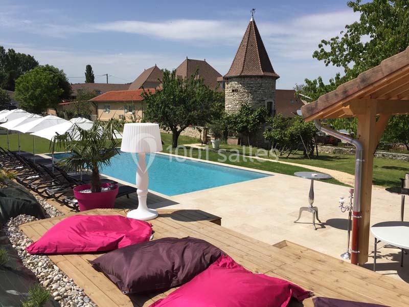 Piscine extérieure entourée de transats, parasols, coussins colorés et vue sur un bâtiment en pierre avec une tour.