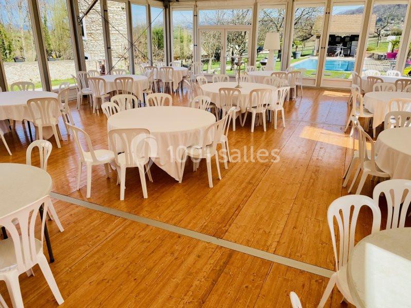 Salle lumineuse avec tables rondes recouvertes de nappes blanches et chaises en plastique, vue sur un jardin et une piscine.