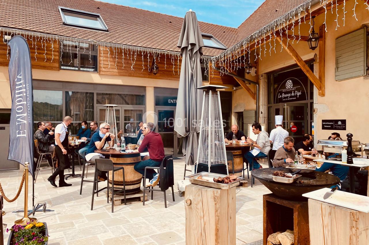 Des personnes déjeunent en terrasse dans une cour ensoleillée, entourée de bâtiments avec des décorations lumineuses.