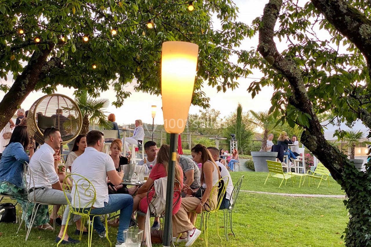 Groupe de personnes assises autour de tables dans un jardin, éclairé par des lampes et entouré de verdure.