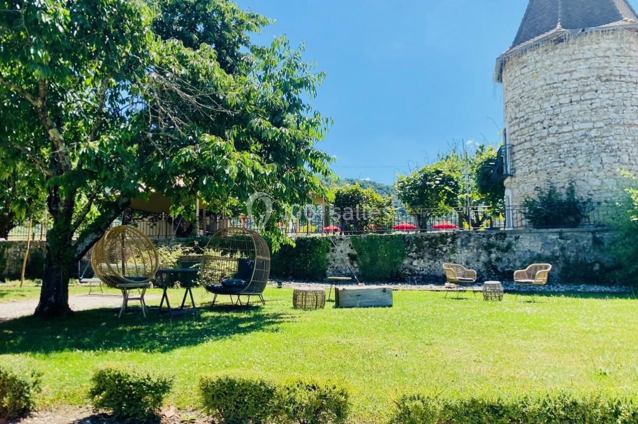 Jardin verdoyant avec chaises en rotin, arbres ombragés et une tour en pierre en arrière-plan.