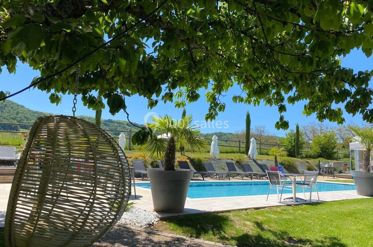Piscine extérieure entourée de chaises longues, parasols et végétation, avec un fauteuil suspendu au premier plan.