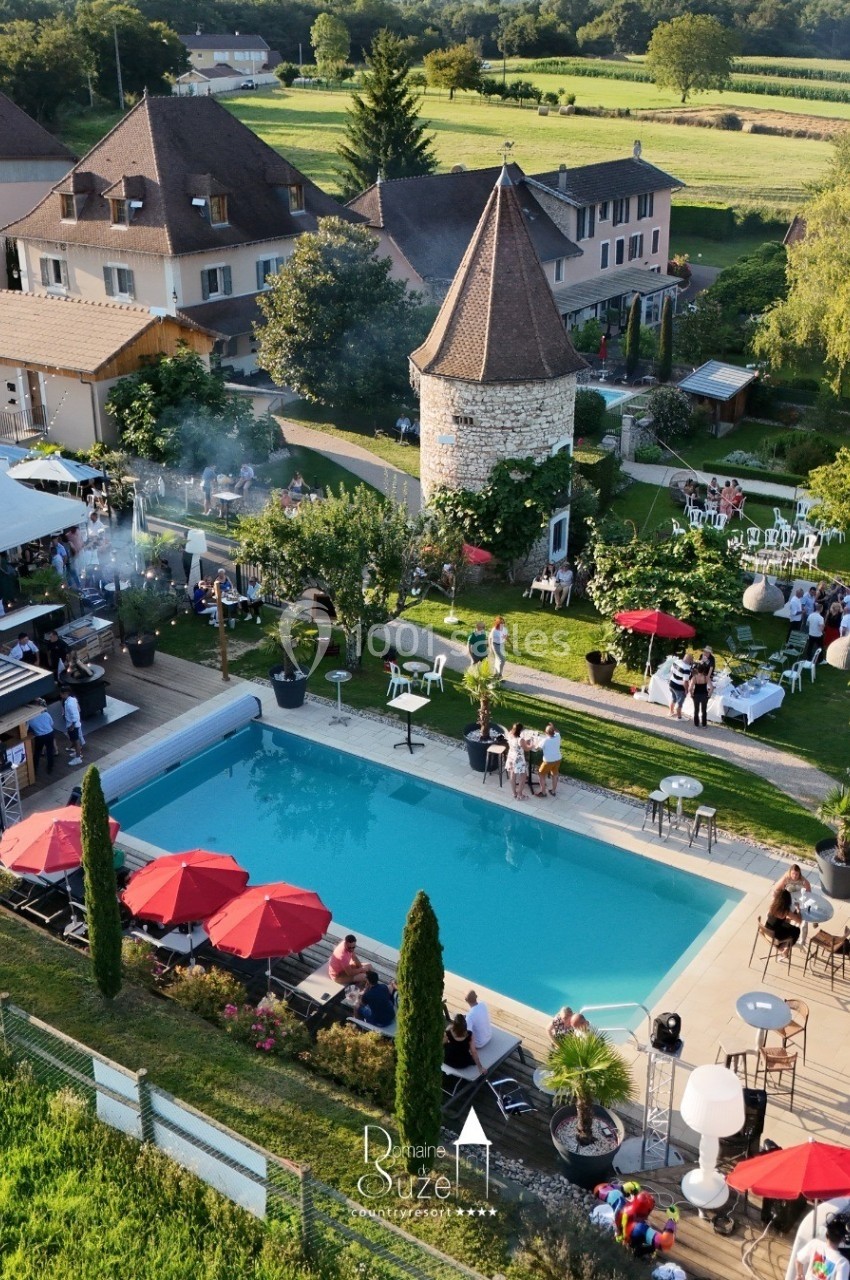 Vue aérienne d'une propriété avec piscine, terrasse, parasols rouges, et bâtiments entourés de verdure.