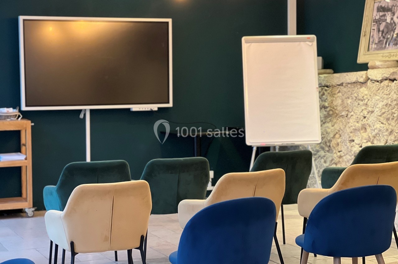 Salle de réunion avec chaises colorées, tableau blanc, écran et mur en pierre apparente.