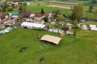 Vue aérienne d'un domaine rural avec des bâtiments, une tente beige, des espaces verts et des participants dispersés.