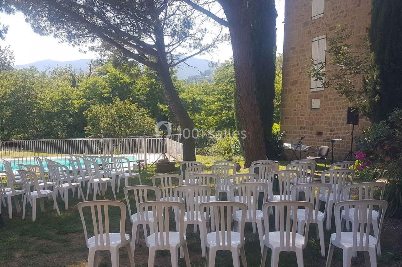 Chaises blanches disposées en extérieur près d'une piscine clôturée, entourées d'arbres et d'un bâtiment en pierre.