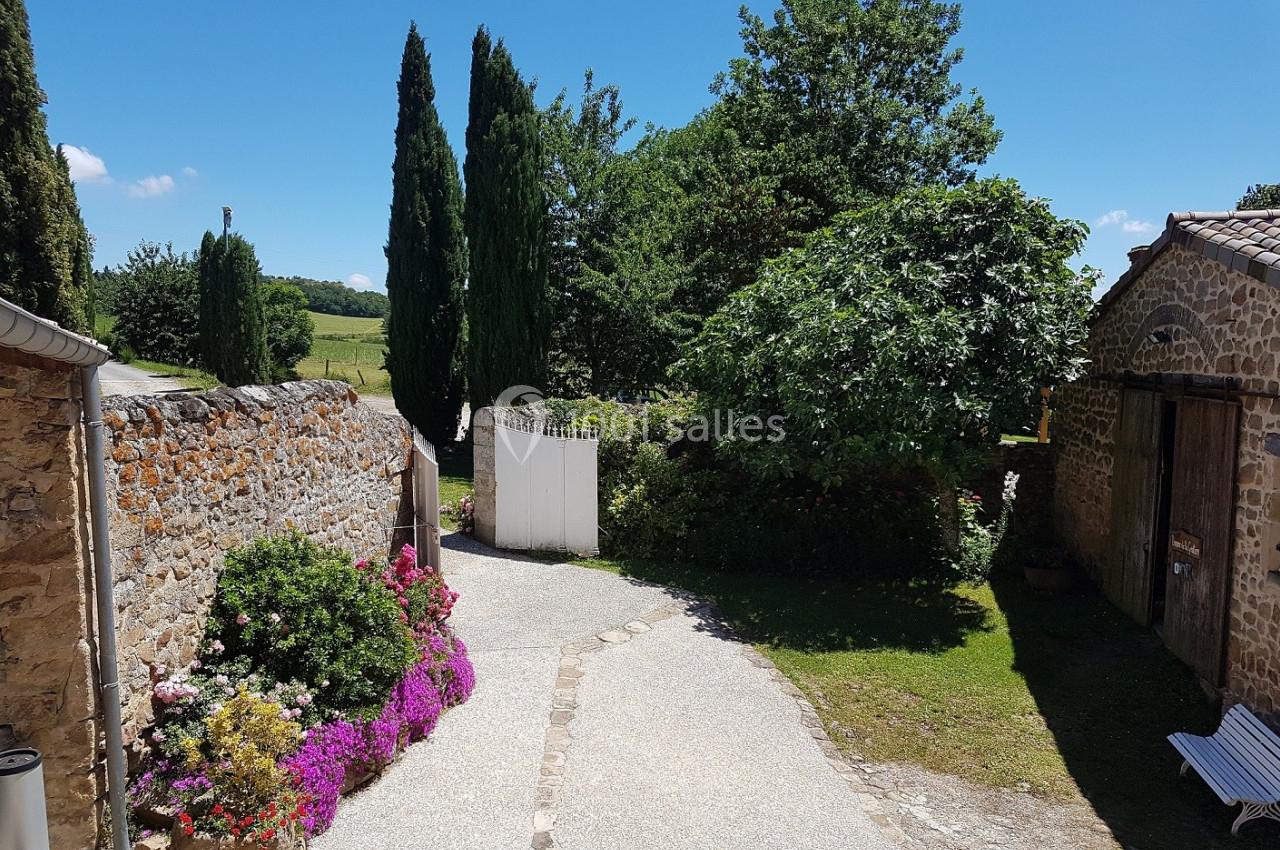 Allée pavée bordée de fleurs et de murs en pierre, menant à un portail blanc, entourée de verdure et d'arbres.