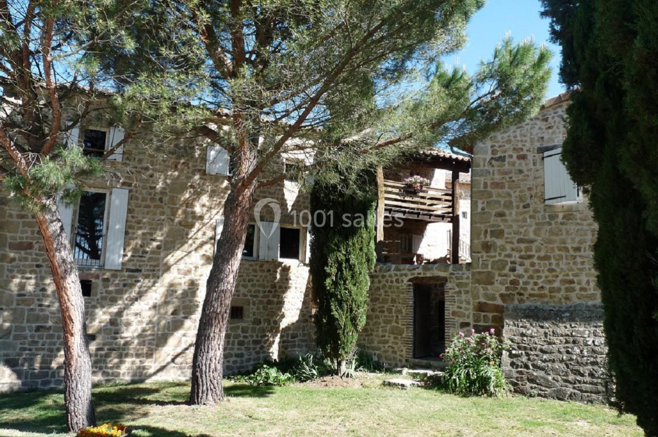 Façade en pierre d'une maison entourée d'arbres et d'un jardin verdoyant sous un ciel bleu.