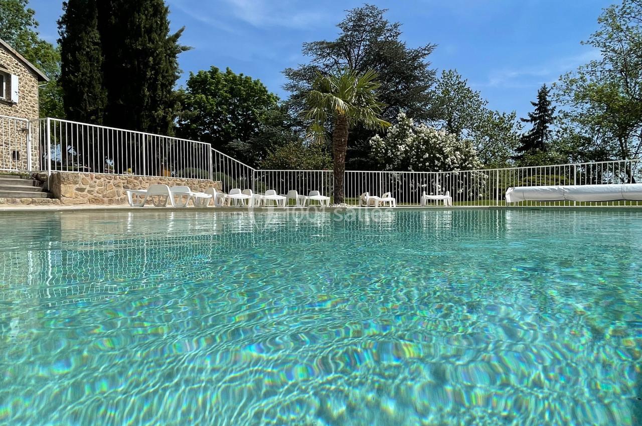 Piscine extérieure entourée de chaises longues blanches, bordée par une clôture et des arbres sous un ciel bleu.