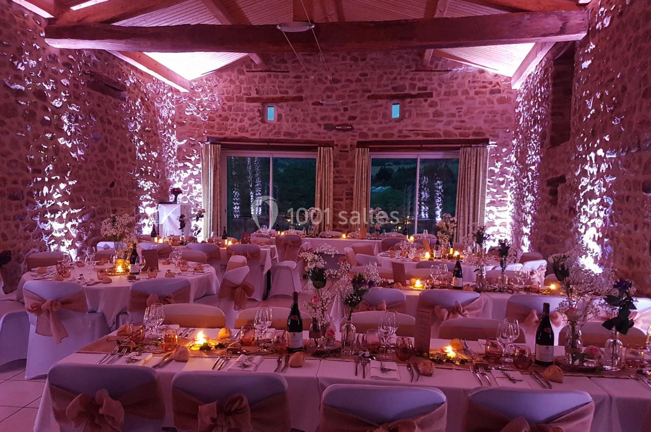 Salle en pierre décorée pour un mariage, avec tables dressées, nappes blanches, rubans beige et éclairage tamisé.