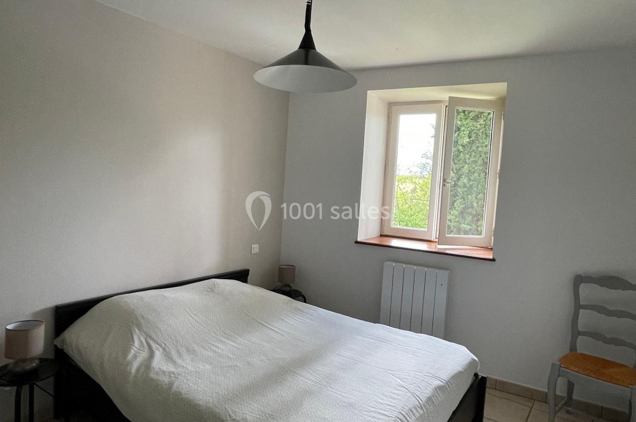 Chambre lumineuse avec un lit double, une suspension noire, une chaise grise et une fenêtre donnant sur un jardin.