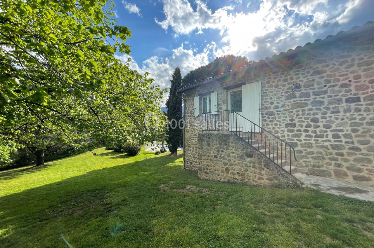 Maison en pierre avec escalier extérieur, entourée d'arbres et d'une pelouse sous un ciel ensoleillé avec quelques nuages.