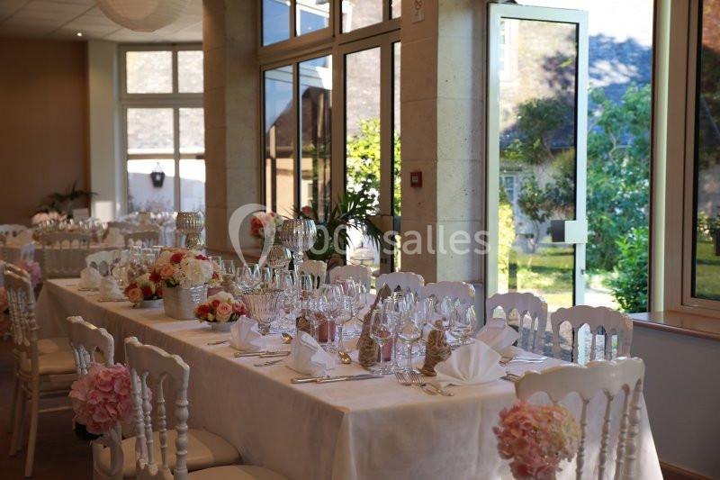 Salle de réception lumineuse avec tables décorées de fleurs, vaisselle élégante et vue sur un jardin verdoyant.
