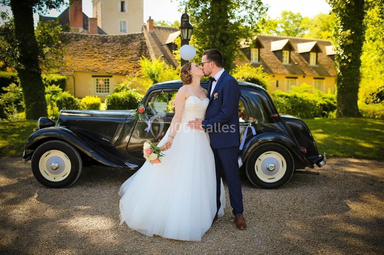 Un couple en tenue de mariage s'embrasse devant une voiture ancienne, dans un cadre verdoyant et ensoleillé.