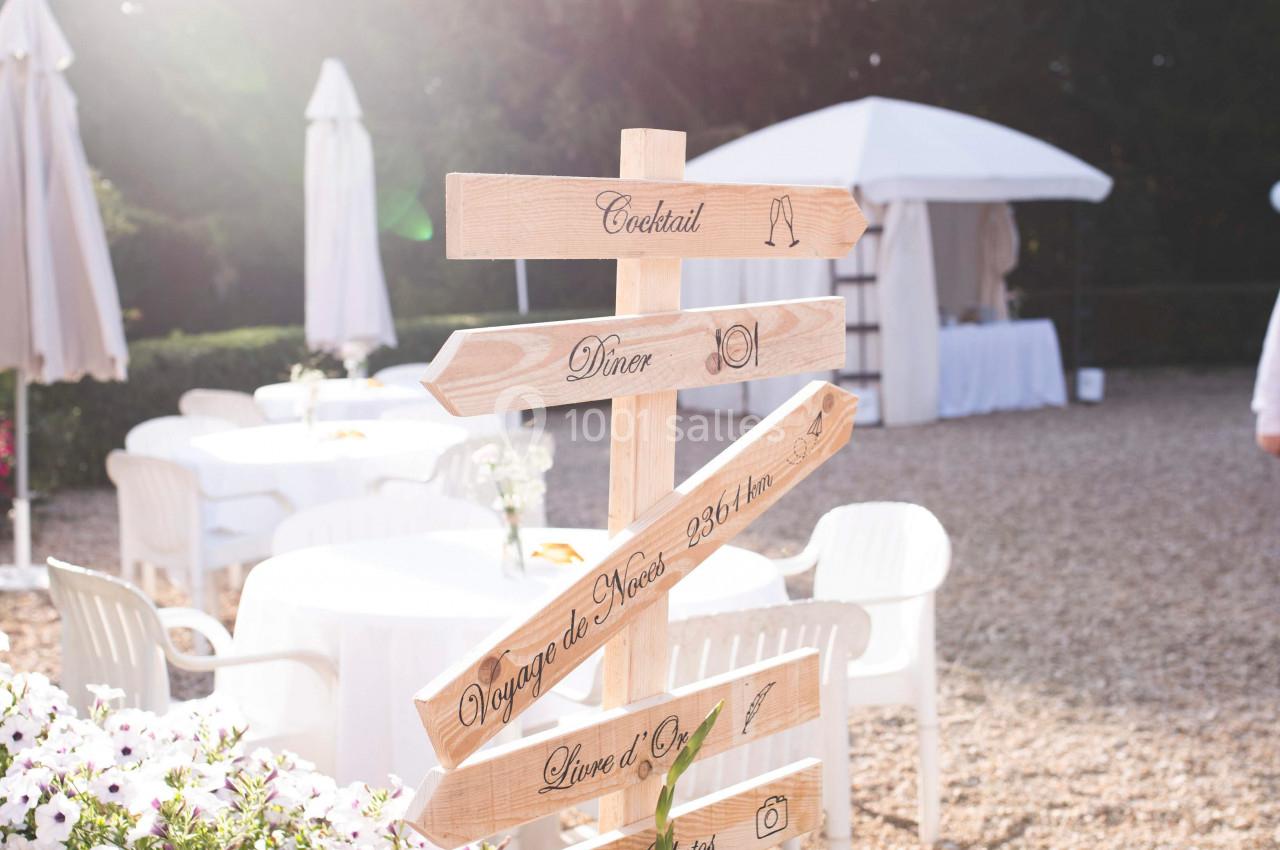 Panneau en bois indiquant différentes directions lors d'un événement en extérieur, entouré de fleurs et de mobilier blanc.