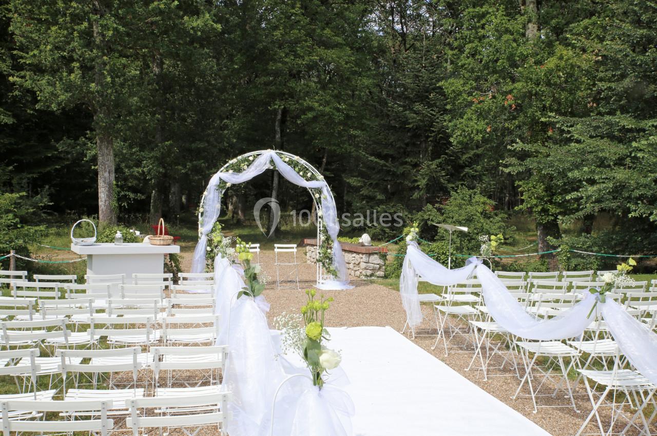 Allée décorée de voilages blancs menant à une arche fleurie pour une cérémonie en plein air dans un cadre boisé.