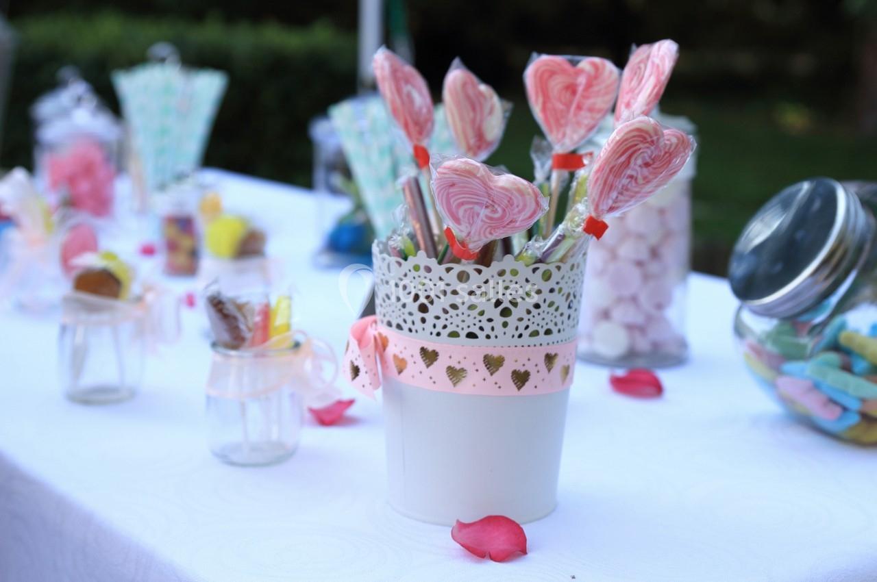 Des sucettes en forme de cœur dans un pot décoré, entourées de bonbons variés sur une table blanche en extérieur.