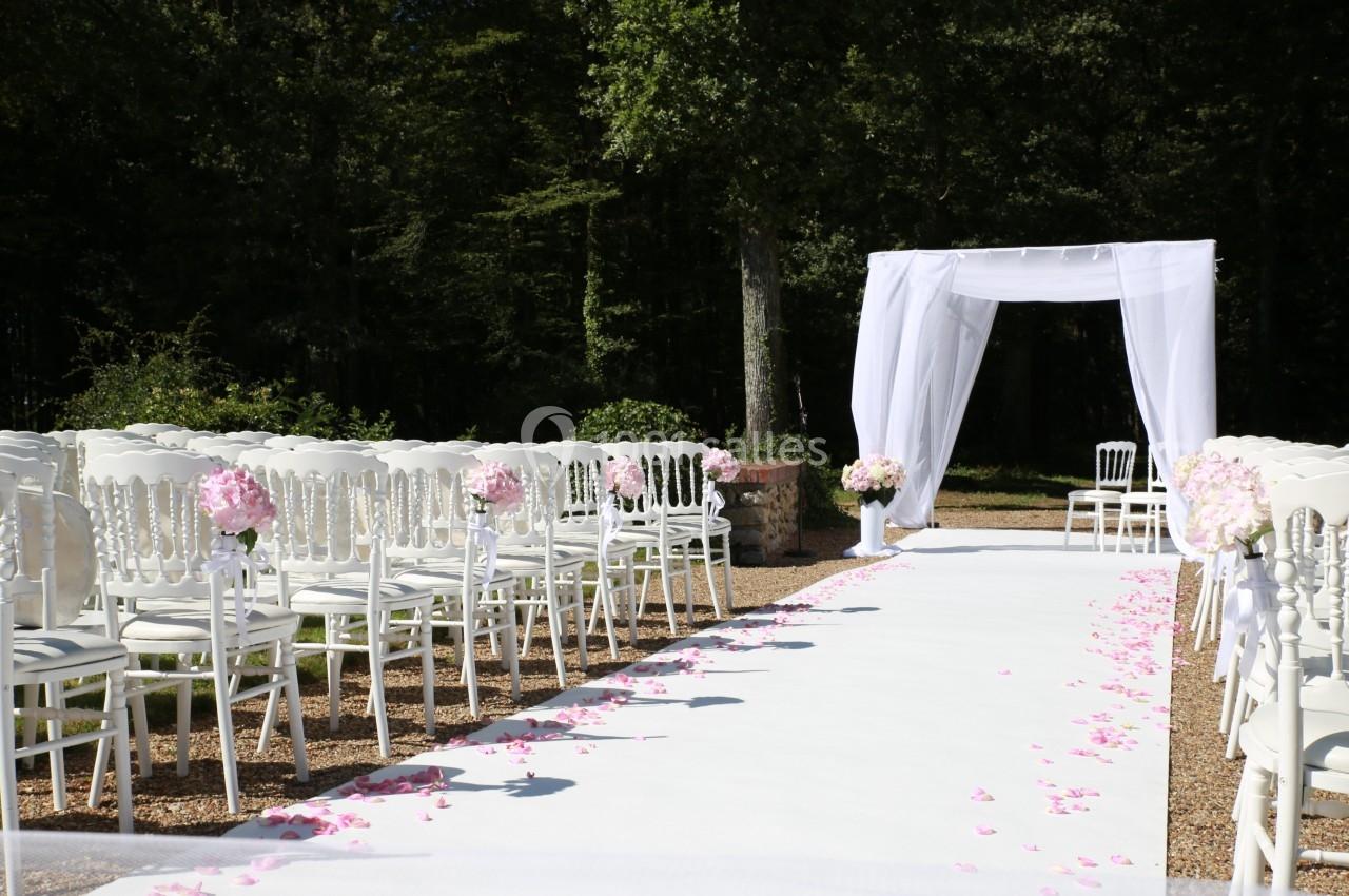 Allée centrale décorée de pétales roses, entourée de chaises blanches, menant à une arche blanche en extérieur.