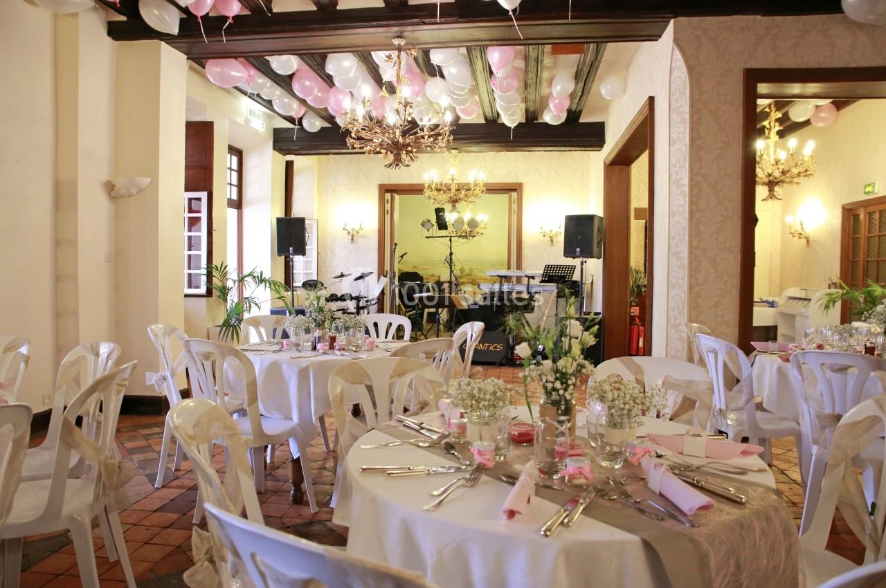 Salle de réception décorée pour un événement, avec tables rondes dressées, chaises blanches et ballons suspendus.