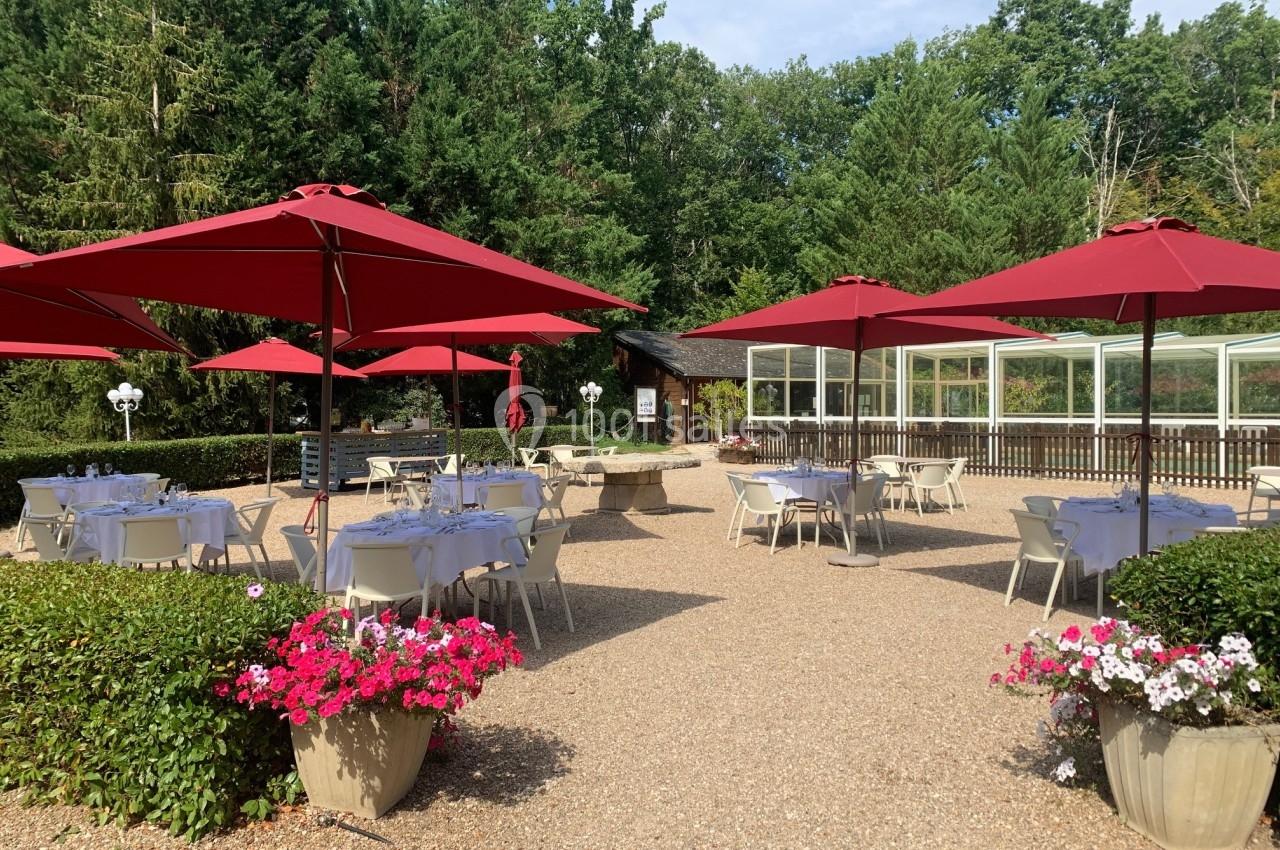 Terrasse extérieure avec tables et chaises blanches sous des parasols rouges, entourée de verdure et de fleurs.