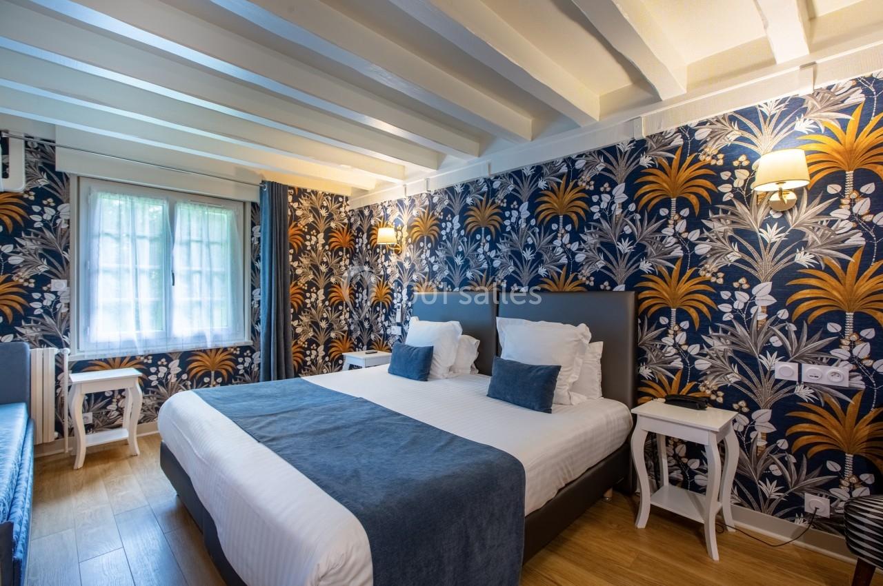 Chambre lumineuse avec lit double, murs décorés de papier peint floral, parquet clair et fenêtres avec rideaux.