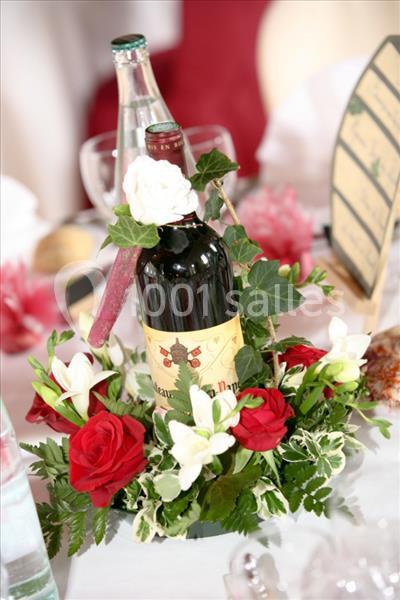 Bouteille de vin rouge décorée de fleurs rouges et blanches, entourée de feuillage sur une table dressée.