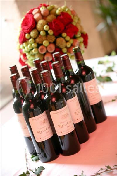 Bouteilles de vin rouge alignées sur une table, avec un bouquet de fleurs et de fruits en arrière-plan.