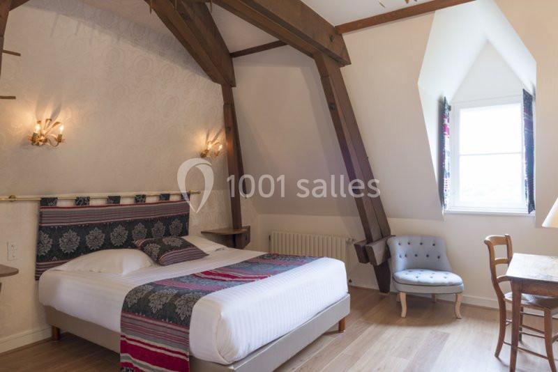 Chambre mansardée avec poutres apparentes, lit double, fauteuil, bureau en bois et fenêtre laissant entrer la lumière…