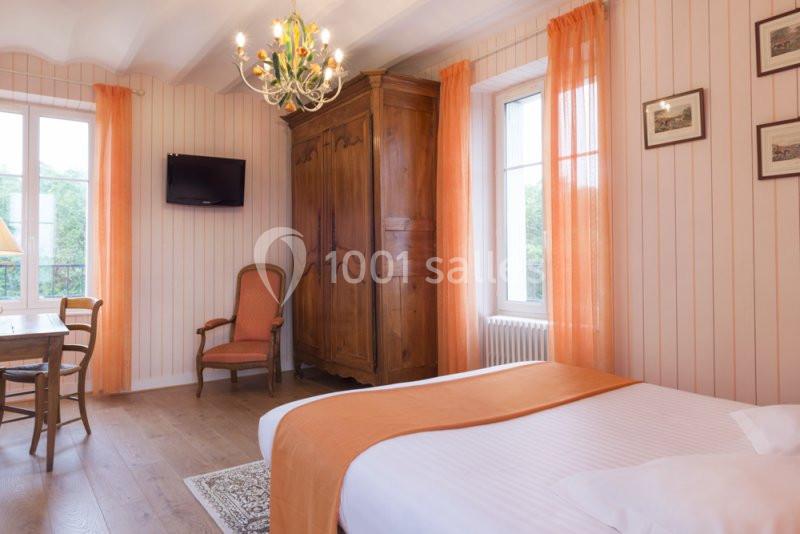 Chambre lumineuse avec lit double, armoire en bois, fauteuil, télévision murale et rideaux orange.