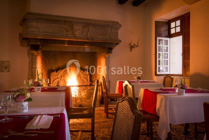Salle de restaurant chaleureuse avec cheminée allumée, tables dressées avec nappes blanches et rouges, lumière tamisée.