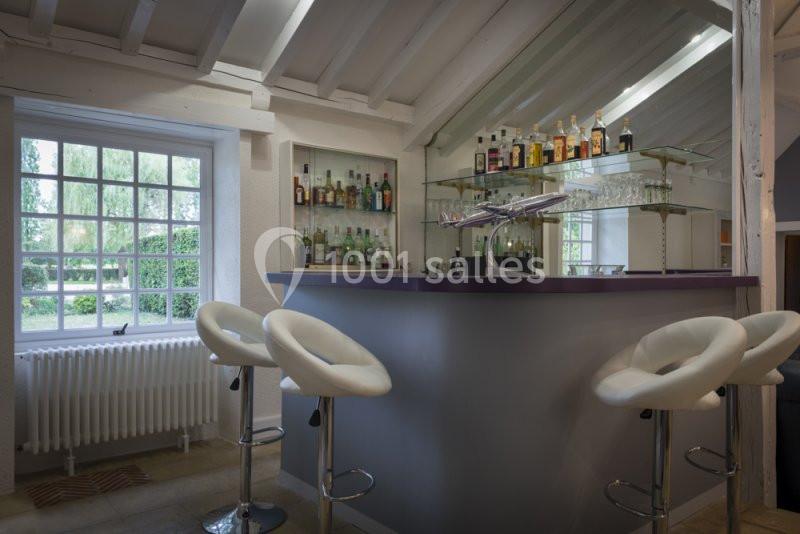 Bar moderne avec comptoir violet, tabourets blancs et étagères de bouteilles, situé dans une pièce lumineuse avec fenêtre.