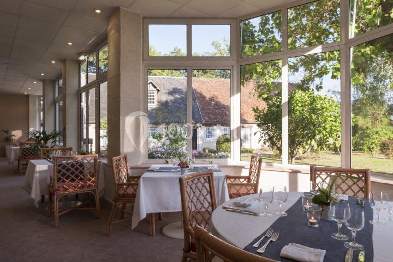 Salle de restaurant lumineuse avec grandes baies vitrées donnant sur un jardin arboré et des bâtiments en arrière-plan.