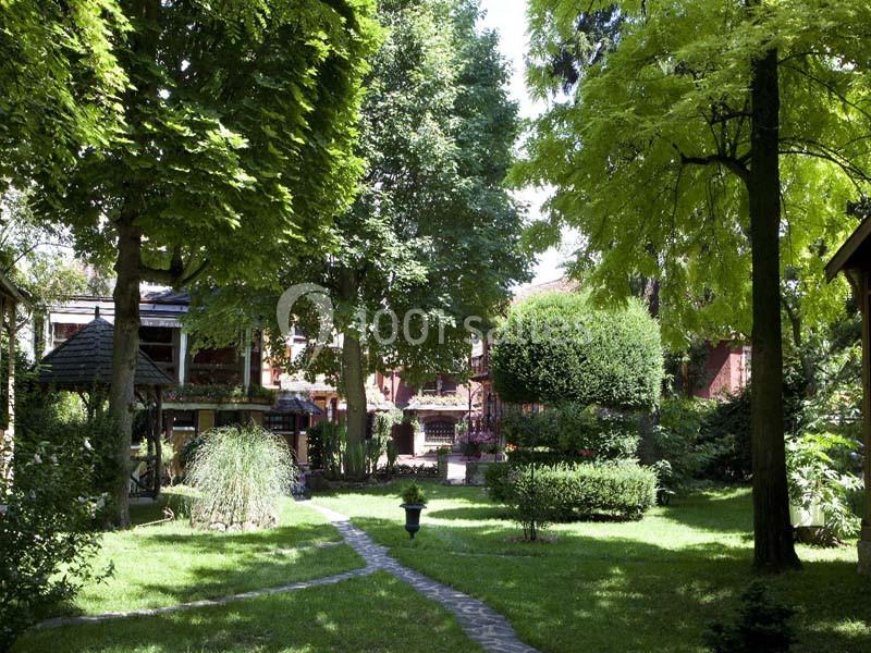 Jardin verdoyant avec allées pavées, arbres ombrageants et bâtiments en briques rouges en arrière-plan.