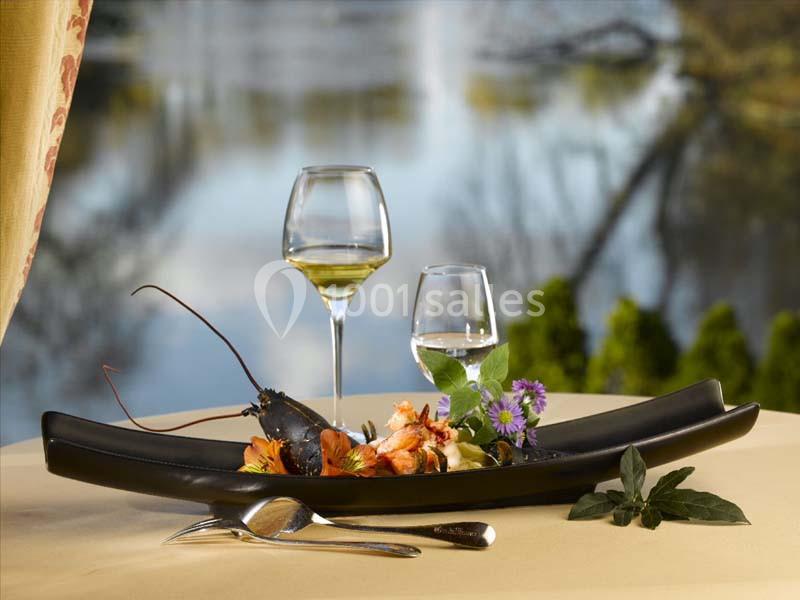 Assiette gastronomique avec homard et fleurs comestibles, accompagnée de deux verres de vin, sur une table près d'un lac.