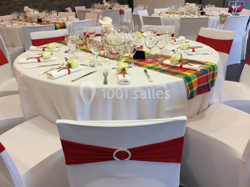 Table ronde décorée pour un événement, avec nappes blanches, chemins de table colorés et chaises ornées de rubans rouges.