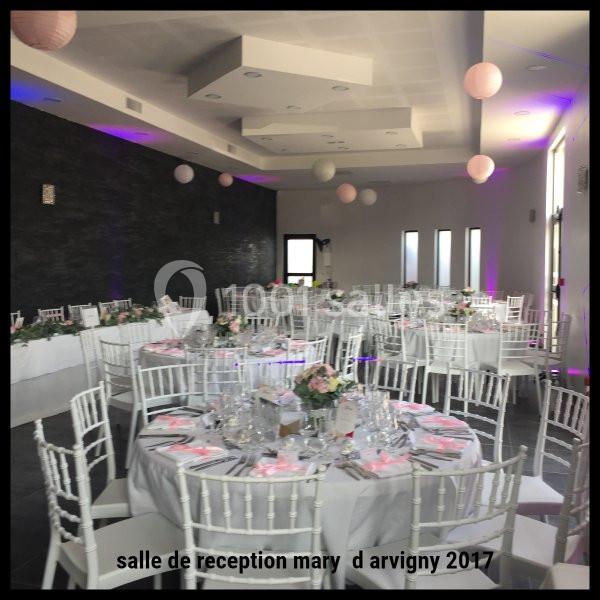 Salle de réception décorée avec des tables rondes, nappes blanches, chaises blanches et éclairage tamisé.