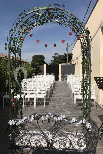 Arche métallique décorée de feuillage et de fleurs blanches, devant des rangées de chaises blanches en extérieur.