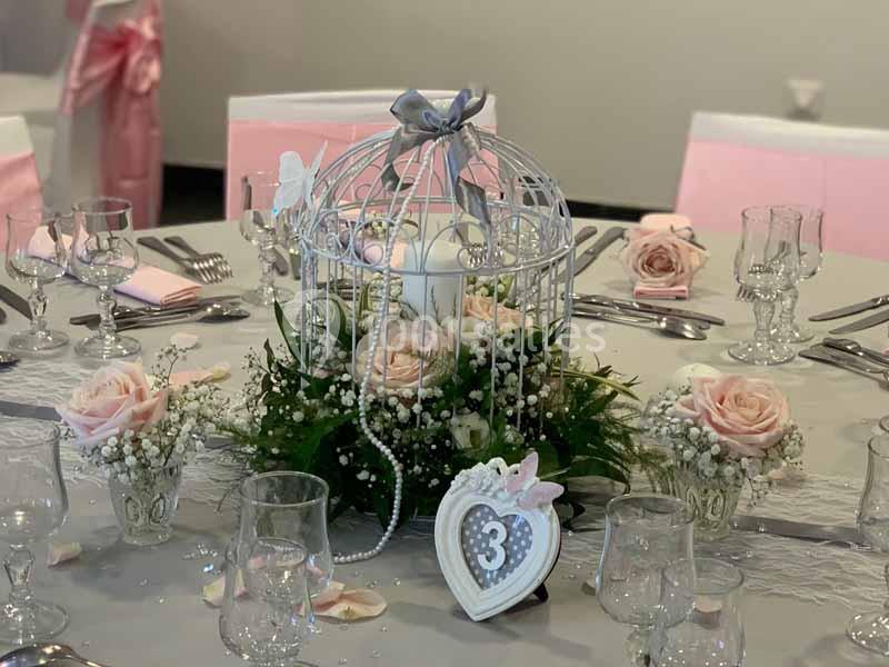 Centre de table avec cage décorative, roses roses, perles et bougies, sur une nappe grise entourée de verres et couverts.