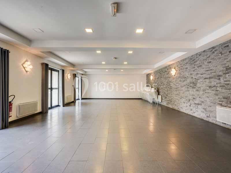 Salle vide avec sol en carrelage, murs clairs dont un en pierre, éclairage au plafond et chaises empilées dans un coin.