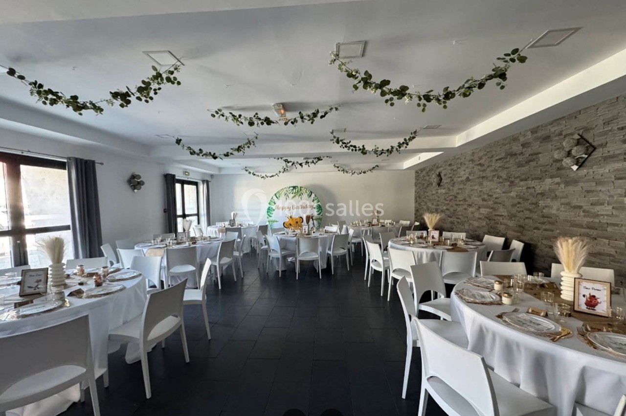 Salle de réception décorée avec des tables rondes, nappes blanches, guirlandes végétales et un mur en pierre.