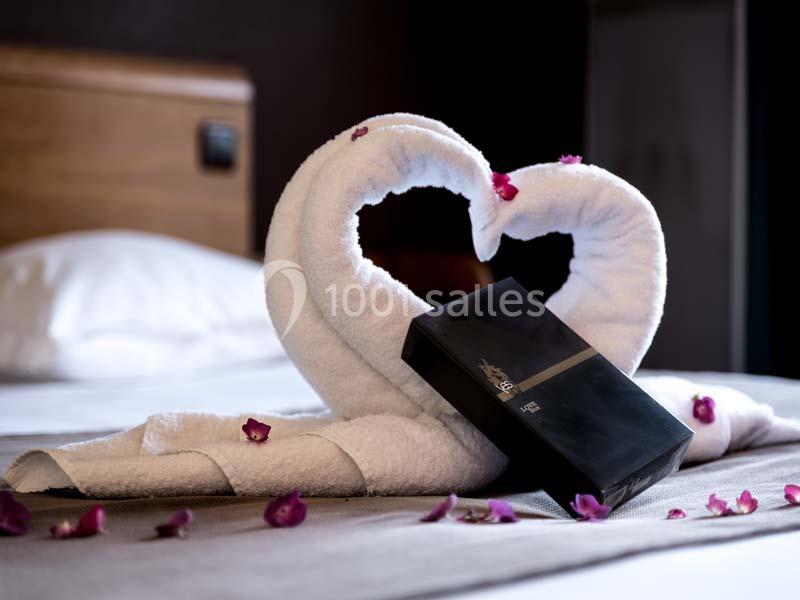 Serviettes pliées en forme de cœur sur un lit, décorées de pétales de fleurs et accompagnées d'une boîte noire.