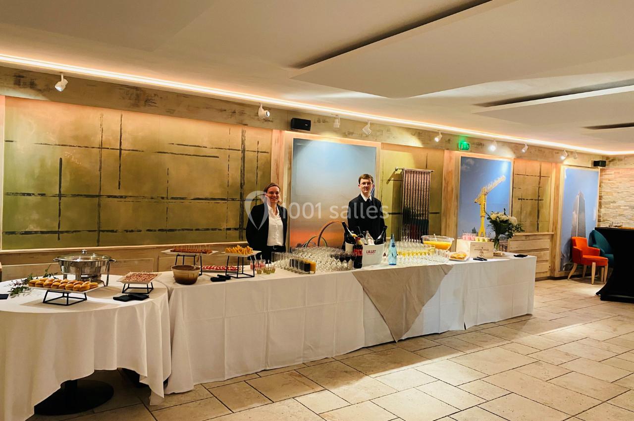 Deux personnes en tenue professionnelle derrière un buffet dressé avec des plats et boissons dans une salle lumineuse.