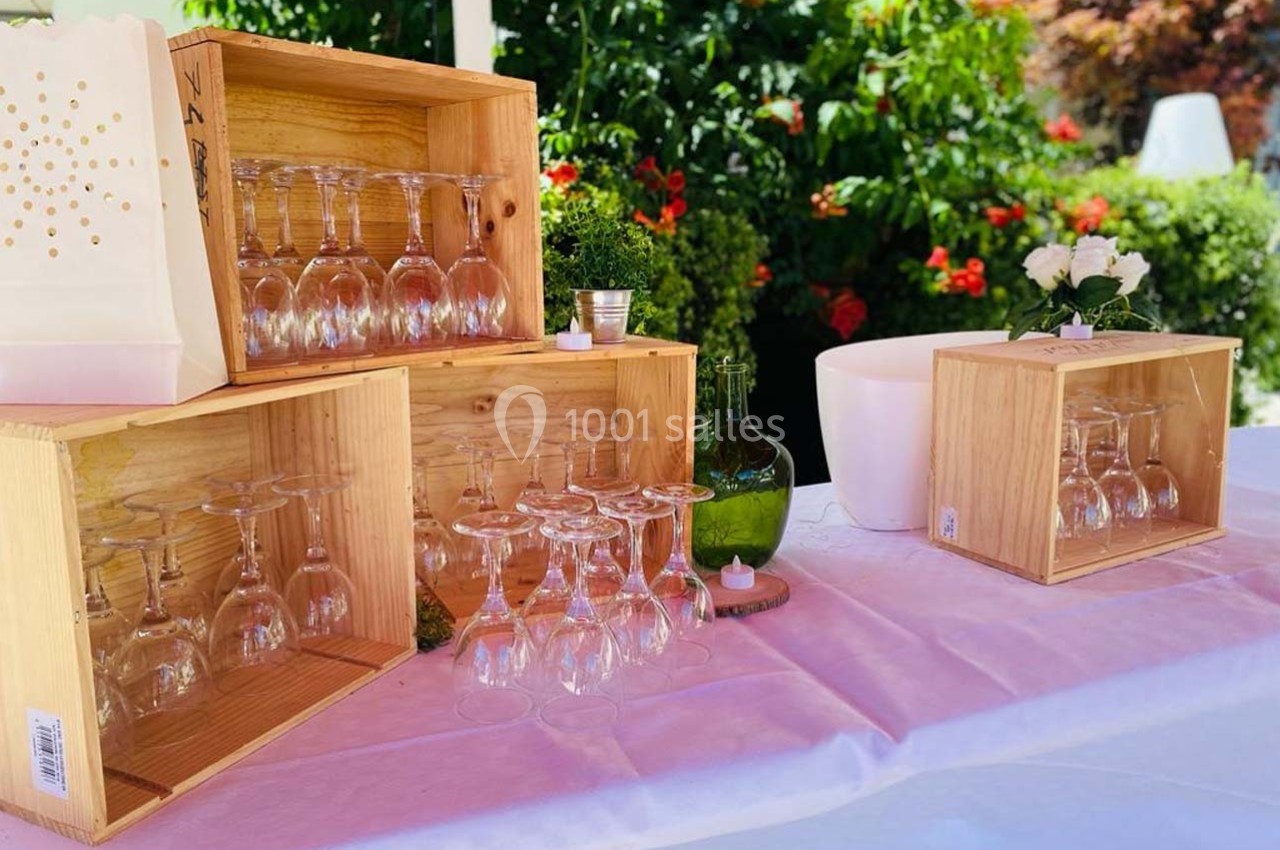 Verres à pied disposés dans des caisses en bois sur une table blanche, avec des plantes en arrière-plan.